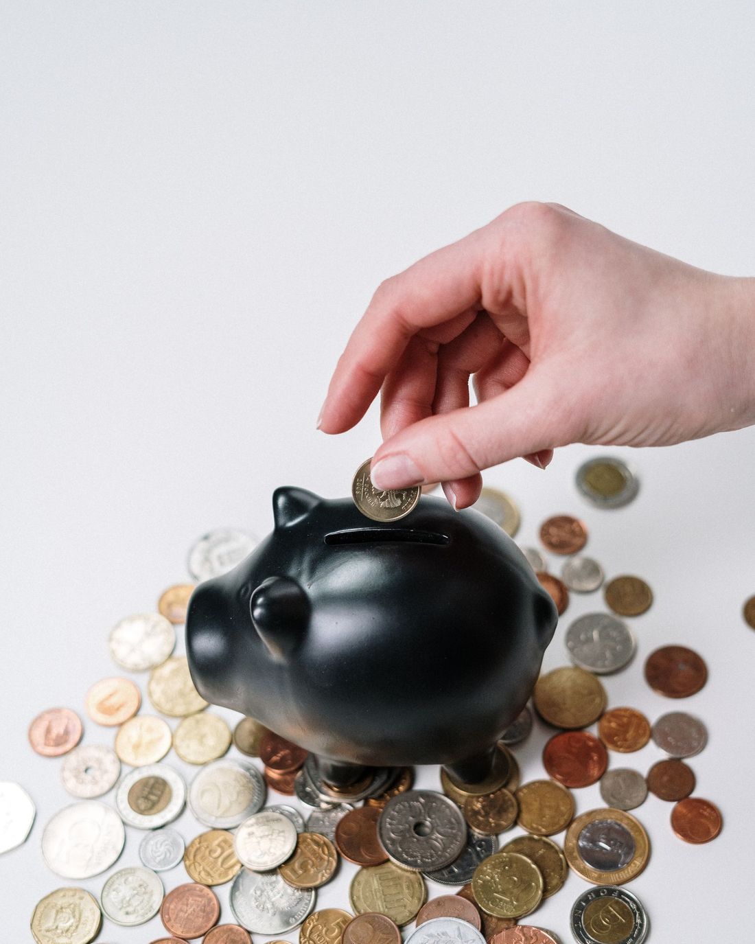 A hand dropping a coin into a piggy bank surrounded by coins